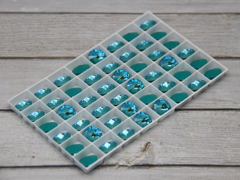 Pu&ograve; includere: Un vassoio bianco pieno di strass color acquamarina e pietre ovali color teal. Il vassoio &egrave; posto su uno sfondo chiaro con venature del legno, creando un contrasto tra le gemme luminose e lo sfondo neutro.