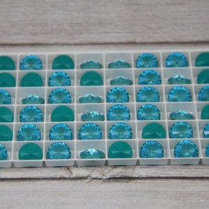 Pu&ograve; includere: Un vassoio bianco pieno di strass di cristallo turchese e verde acqua. Gli strass sono disposti a griglia, con forme a semicerchio e ovali. I cristalli hanno un design sfaccettato, che riflette la luce. Il vassoio &egrave; su una superficie di legno chiaro.