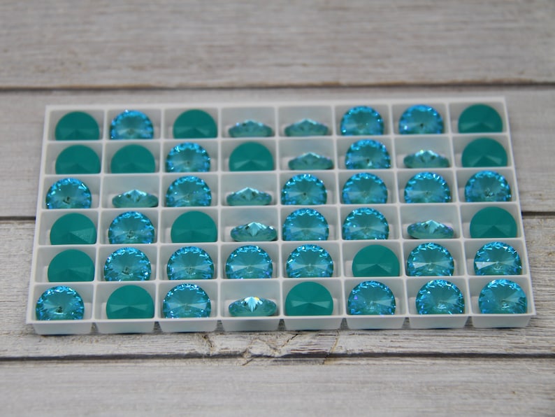 Pu&ograve; includere: Un vassoio bianco pieno di strass di cristallo turchese e verde acqua. Gli strass sono disposti a griglia, con forme a semicerchio e ovali. I cristalli hanno un design sfaccettato, che riflette la luce. Il vassoio &egrave; su una superficie di legno chiaro.
