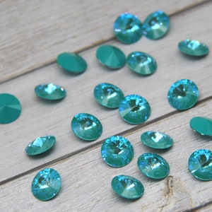 Pu&ograve; includere: Una collezione di strass in cristallo sfaccettato color verde acqua. I cristalli sono di forma rotonda e ovale, con una finitura brillante e iridescente. Alcuni cristalli sono piatti, mentre altri hanno il retro a punta. I cristalli sono sparsi su una superficie di legno grigio chiaro.