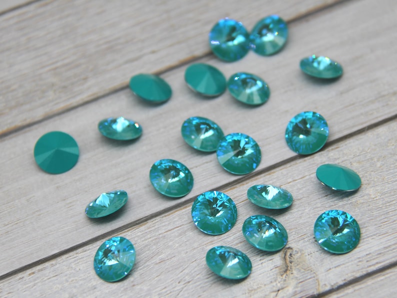 Pu&ograve; includere: Una collezione di strass in cristallo sfaccettato color verde acqua. I cristalli sono di forma rotonda e ovale, con una finitura brillante e iridescente. Alcuni cristalli sono piatti, mentre altri hanno il retro a punta. I cristalli sono sparsi su una superficie di legno grigio chiaro.