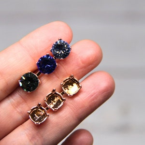 May include: Close-up of two sets of jewelry components. One set features three round, faceted gemstones in shades of green, blue, and purple. The other set is gold-toned with three square settings. The components are held in a hand.