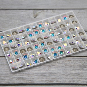 May include: A white plastic tray filled with iridescent blue and clear rhinestones. The rhinestones are arranged in a grid pattern.