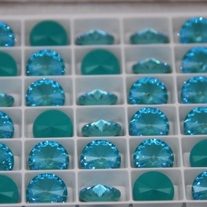 Pu&ograve; includere: Un vassoio pieno di strass di cristallo di vari colori turchese e verde acqua. Gli strass sono disposti in una griglia, alcuni a forma di semicerchio e altri con un design sfaccettato a ventaglio. I cristalli sono esposti in un organizer di plastica bianca.