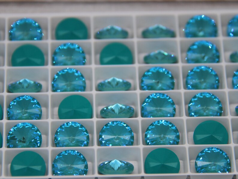 Pu&ograve; includere: Un vassoio pieno di strass di cristallo di vari colori turchese e verde acqua. Gli strass sono disposti in una griglia, alcuni a forma di semicerchio e altri con un design sfaccettato a ventaglio. I cristalli sono esposti in un organizer di plastica bianca.