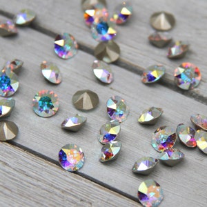 May include: Assortment of iridescent glass rhinestones in various shades of blue, green, and purple. The rhinestones are scattered on a light grey wooden surface.