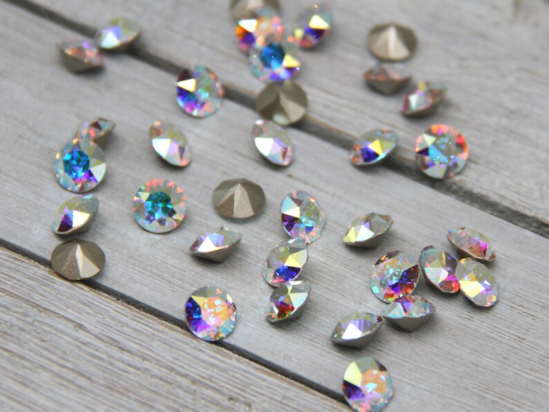 May include: Assortment of iridescent glass rhinestones in various shades of blue, green, and purple. The rhinestones are scattered on a light grey wooden surface.