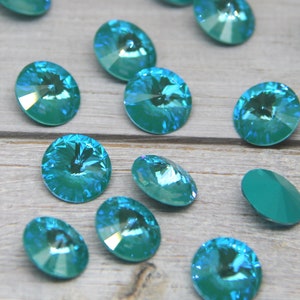Pu&ograve; includere: Una collezione di cristalli ovali color teal con superfici sfaccettate. I cristalli sono sparsi su una superficie di legno chiaro, riflettendo la luce e creando un effetto scintillante. Questi cristalli decorativi sono adatti per la creazione di gioielli e l'artigianato.