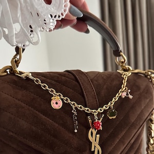 May include: A brown suede YSL handbag with a gold chain strap and a charm featuring a ladybug, donut, and other small charms. The charm also has the word "mama" on it.