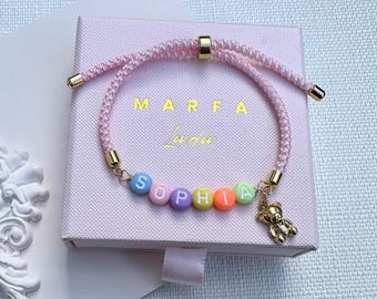May include: A pink cord bracelet featuring colorful letter beads spelling the name SOPHIA, accented with gold-toned metal beads and a small gold-toned teddy bear charm. The bracelet is displayed on a light pink square box with the text MARFA LULU.