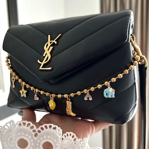 Pode incluir: Bolsa de ombro acolchoada preta com um logotipo YSL dourado e uma alça de corrente dourada com pingentes coloridos. A corrente mede 30,5 cm de comprimento.