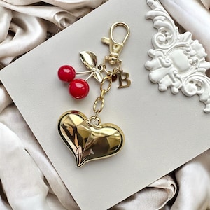 Puede incluir: Un llavero dorado con un dije en forma de corazón, detalles de cerezas rojas y un dije con la letra "B". El llavero tiene un cierre de mosquetón. El dije de corazón es facetado.