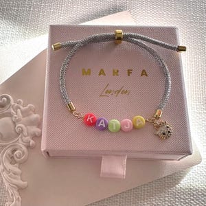 May include: A silver bracelet with the name "KATJA" spelt out in colourful letter beads. The bracelet has gold-coloured accents and a charm. The bracelet is displayed on a pink box with the text "MARFA London".
