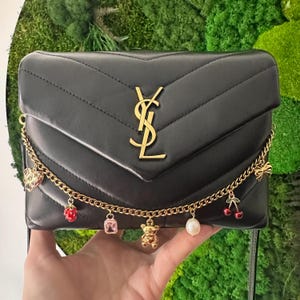 May include: Black quilted leather shoulder bag with a gold chain strap and charms. The charms include a teddy bear, a pearl, a cherry, a pink gem, and a red strawberry.