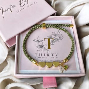May include: A green cord bracelet with gold accents and heart charms, presented in a pink box. The box features the text "THIRTY HAPPY 30TH BIRTHDAY! HEARTS FOR THREE DECADES" and the letter "T". The bracelet includes a charm with the letter "A".