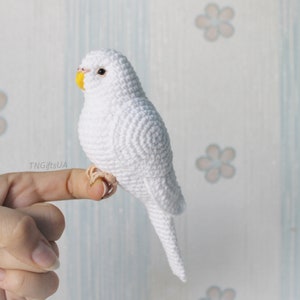 Puede incluir: Un periquito blanco de crochet con un pico amarillo y ojos negros. El periquito está posado en un dedo.