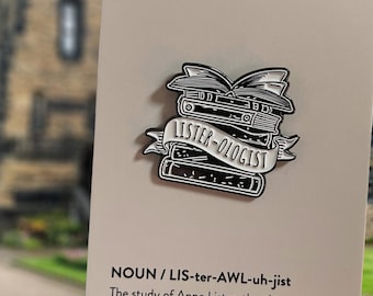 May include: A black and white enamel pin depicting a stack of books with an open book on top. The pin has a banner that reads "LISTER-OLOGIST". The background is a blurred image of a building and a card with text.