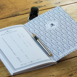 Puede incluir: Un libro abierto con el texto "ANNE LISTER'S CODE" y una pluma estilográfica plateada y dorada sobre una superficie de madera. Un pequeño frasco de tinta negra está junto al libro. La portada del libro tiene un patrón azul y blanco.