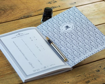 May include: An open book with the text "ANNE LISTER'S CODE" and a silver and gold fountain pen on a wooden surface. A small bottle of black ink sits next to the book. The book's cover has a blue and white pattern.
