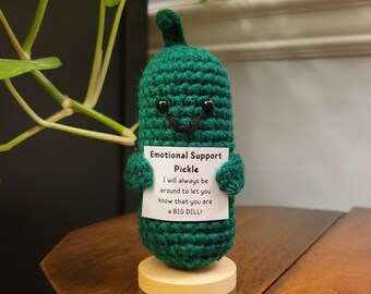 May include: A green, crocheted pickle figure with a smiling face, holding a sign that reads "Emotional Support Pickle." The sign says "I will always be around to let you know that you are a BIG DILL!" The pickle is on a small wooden base.