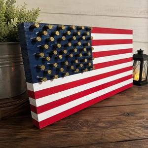 May include: A rectangular wooden wall art piece painted to resemble the American flag, featuring red and white horizontal stripes and a blue canton. The blue section is adorned with rows of metallic brass-colored bullet casings as stars.
