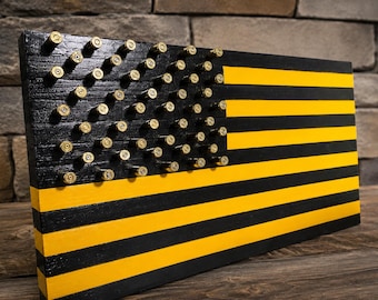 May include: A rectangular wooden wall art piece designed as a flag with alternating horizontal black and yellow stripes. The canton area is painted black and features a grid of metallic brass bullet casings arranged to represent the stars.