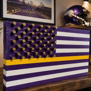 May include: A rectangular wooden wall art piece designed as a flag with horizontal purple and white stripes. The canton features rows of metallic gold-colored bullet casings. A single horizontal yellow stripe runs across the lower section.
