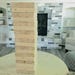 May include: A freestanding, light-colored wooden tower game, similar to Jenga, is shown.  The blocks are rectangular and stacked vertically, creating a tall structure. The blocks are light beige or natural wood color. The game is positioned on a light-colored table in front of a fireplace with gray and white brickwork. The background includes a living room with shelves and furniture.