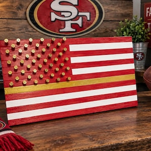 May include: A rectangular wooden wall art piece designed as an American flag with red and white horizontal stripes. The blue field is replaced by red wood slats adorned with brass-colored bullet casings, and one stripe is painted in metallic gold.