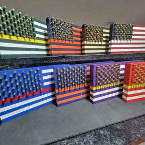 May include: A collection of eight American flag-themed wood plaques with a variety of color schemes. Each plaque features a design of the American flag with a field of stars made of bullet casings.
