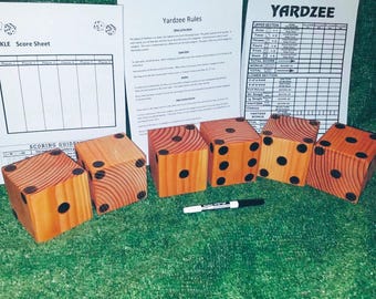 May include: A set of six large wooden dice with black dots, used for the game Yardzee. The dice are on a green surface with a score sheet and rules for the game in the background.