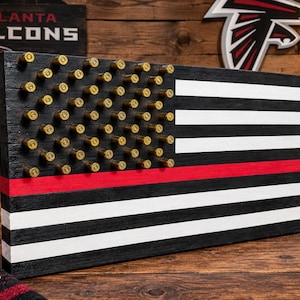 May include: A rectangular wooden wall art piece designed as a flag with alternating black and white horizontal stripes and a single red stripe. The canton area features a grid of brass-colored bullet casings arranged in rows against a black background.