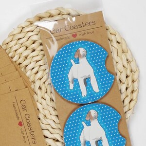 May include: Two car coasters with a white dog illustration on a blue polka dot background. The coasters are packaged with brown cardstock that reads "Car Coasters Handmade with love". A stack of brown cardstock envelopes is also visible.