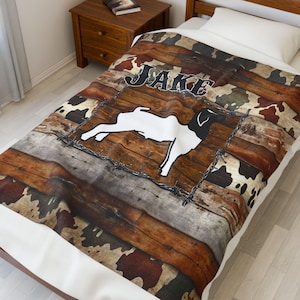 May include: A personalised blanket with a white goat illustration inside a barbed wire frame, set against wooden planks and cowhide patterns. The name "JAKE" is above the goat in a bold font. The blanket is on a bed, with a white pillow.