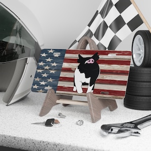 May include: A wooden license plate featuring a black and white pig with a pink nose, standing on a red, white, and blue American flag background. The license plate is on a wooden stand.
