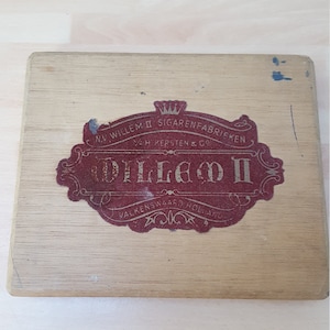 May include: Wooden cigar box with a faded red and gold label. The label features the text "Willem II Sigarenfabrieken" and "Valkenswaard Holland". The box is rectangular and shows signs of wear.