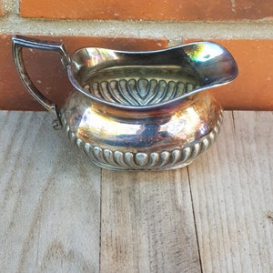 Peut inclure: Une crémier en argent avec un motif décoratif et une poignée. La crémier est posée sur une surface en bois.