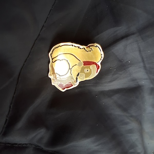 以下が含まれることがあります： 赤いサンタ帽子のついた漫画のリスが描かれた金色のエナメルピン。リスは小さな白い物体を足で持っています。