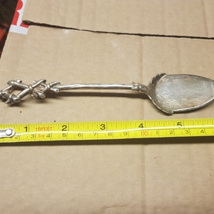 Może przedstawiać: Srebrny łyżka do serwowania z ozdobnym uchwytem i miseczką w kształcie liścia. Łyżka ma około 15 cm długości. Uchwyt ma dekoracyjny wzór. Miseczka ma szczegółowy wzór. Łyżka leży na brązowej powierzchni.