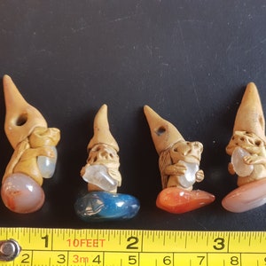 May include: Four small ceramic gnome figurines, each holding a clear crystal and standing on a different colored stone base. The gnomes are wearing brown pointed hats and have detailed facial features.