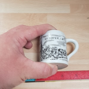 May include: White ceramic mug with a black and white illustration of a city skyline. The mug has a handle and the text "MVNSTER, i.W." printed on the side.