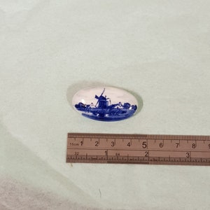 Könnte beinhalten: Eine weiße ovale Keramikfliese mit einem blauen und weißen Bild einer Windmühle in einer holländischen Landschaft. Die Fliese ist etwa 5 cm lang.
