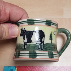 May include: A ceramic mug with a green, white and orange striped design. The mug features a black and white cow illustration with a green field and a small tree in the background.