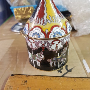 Könnte beinhalten: Eine kleine, farbenfrohe Cloisonné-Schmuckdose mit Karussell-Motiv. Die Dose ist schwarz mit weißen Pferden und einem rot-gelb-weiß gestreiften Dach.