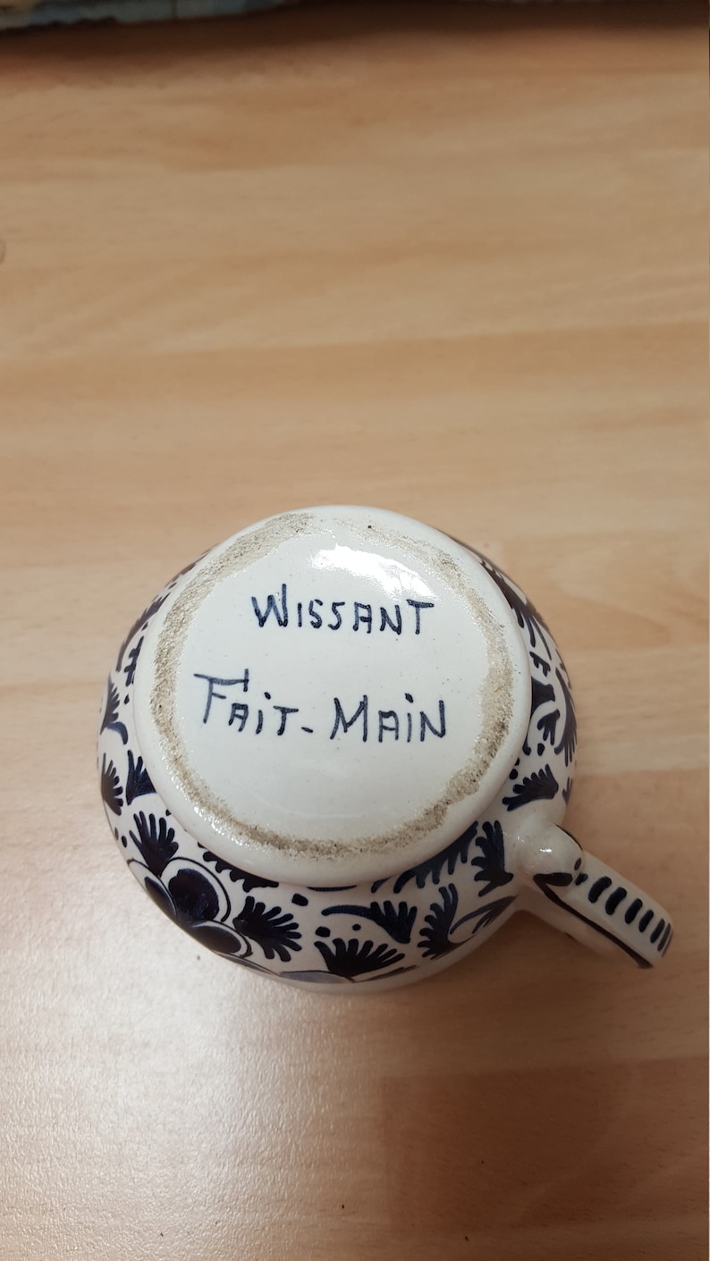 Pu&ograve; includere: Una tazza in ceramica bianca con un motivo floreale blu e bianco. Sul fondo della tazza &egrave; scritto "Wissant Fait-Main".