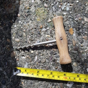 May include: A wooden handled corkscrew with a silver metal spiral. The corkscrew is lying on a gray concrete surface with small pebbles. A yellow measuring tape is partially visible in the bottom of the image.