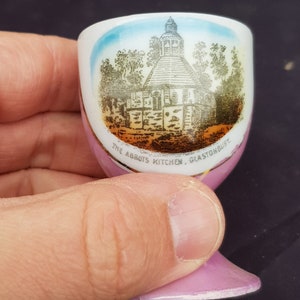 Puede incluir: Un pequeño vaso para huevos de cerámica rosa y blanco con un borde dorado. El vaso presenta una ilustración en blanco y negro de un edificio con el texto "The Abbots Kitchen, Glastonbury" impreso en el lateral.