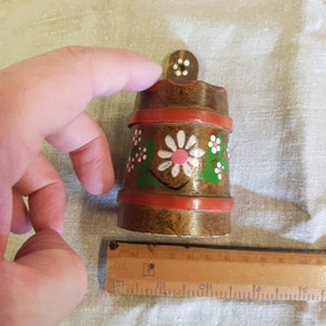 May include: A small wooden container with a red, white, and green floral design. The container has a small wooden lid with a white dot in the center.