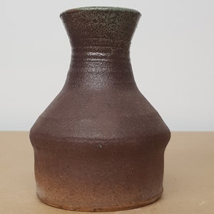 Puede incluir: Un jarrón de cerámica marrón con un esmalte verde en el borde superior. El jarrón tiene un cuerpo redondeado y un cuello estrecho.