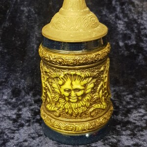 May include: A ceramic beer stein with a green and gold design. The stein features a detailed face of a green man with a beard and a crown on top of the lid.